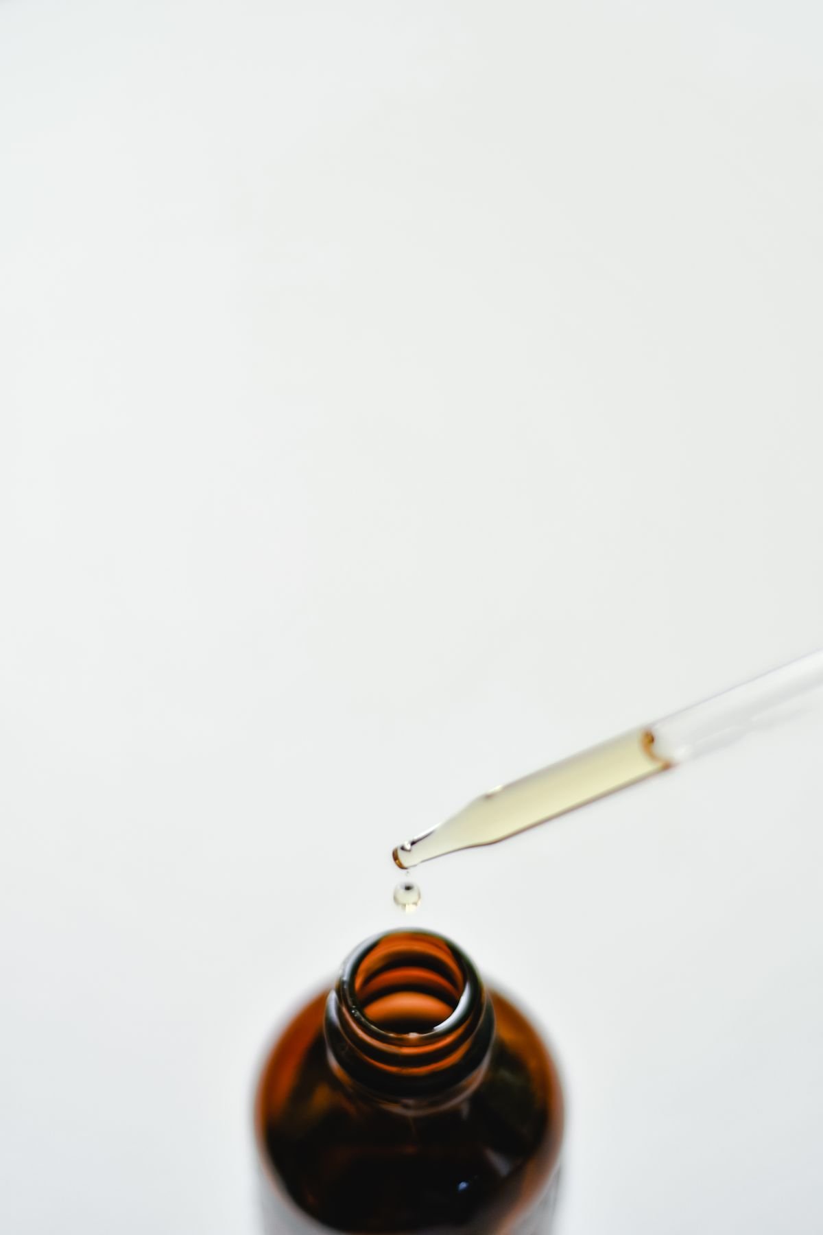 A glass dropper releasing a drop of CBD oil over an open amber bottle on a white background