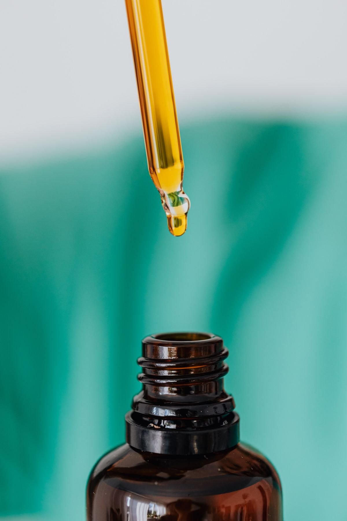 Golden full-spectrum CBD oil dripping from a glass dropper into an amber bottle on a teal background